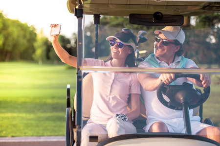 Beautiful couple is smiling while driving a golf cart, girl is taking photos using a smart phoneの写真素材