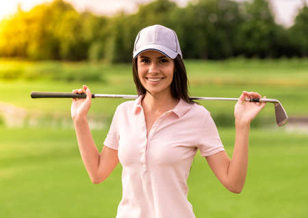 Beautiful young woman is holding a golf club, looking at camera and smiling while standing on golf courseの写真素材