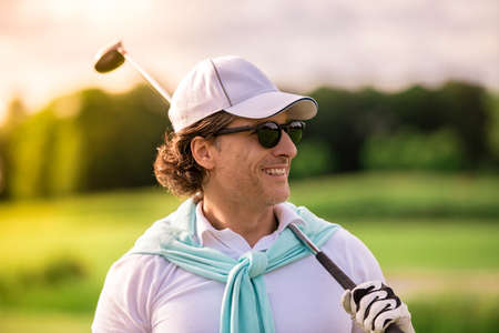 Handsome man is holding a golf club, looking away and smiling while standing on golf courseの写真素材