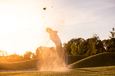Handsome guy is using a golf club while playing golf, beautiful sun shining, splashed of sandの写真素材