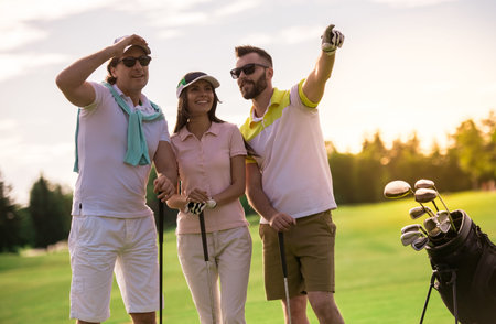 Two men and a woman are holding golf clubs, talking and smiling while standing on golf courseの写真素材