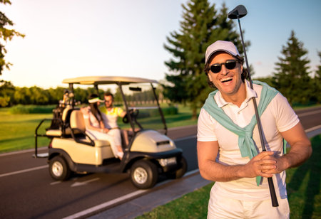 People playing golf. Handsome man in the foreground holding a golf club and smiling, young couple in a golf cartの写真素材
