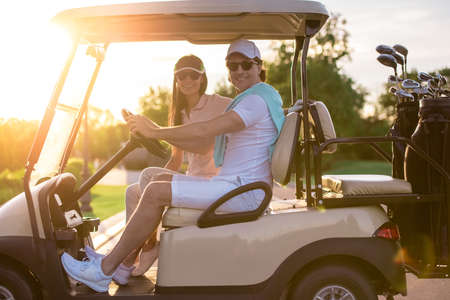 Beautiful couple is looking at camera and smiling while driving a golf cartの写真素材