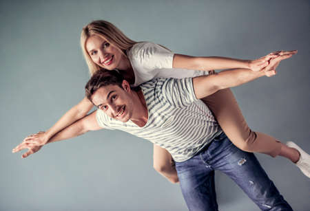Beautiful young couple is having fun, looking at camera and smiling, on gray background, girl is pickabackの写真素材