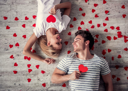 Top view of beautiful young couple holding red paper hearts, looking at each other and smiling while...