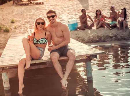 Happy couple is drinking soda water, looking at camera and smiling while sitting on pier at the seaの写真素材
