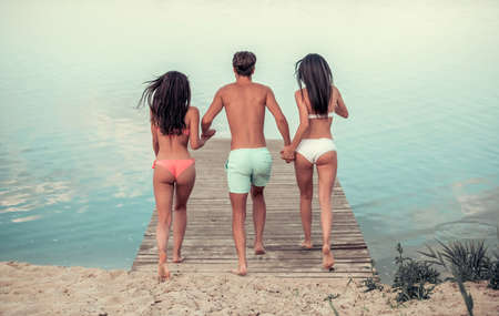 Back view of beautiful girls and guy holding hands and running along the pier ready to jump into the waterの写真素材
