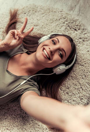 Beautiful woman in casual clothes and headphones is listening to music, showing peace sign and smiling while lying on the floor at homeの写真素材