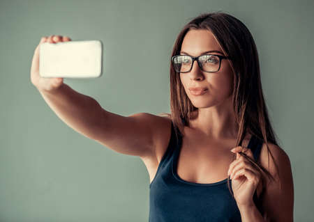 Beautiful woman in casual clothes and eyeglasses is doing selfie using a smart phone, on gray backgroundの写真素材