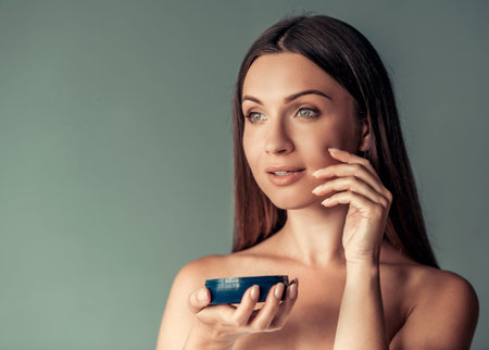 Beautiful woman with bare shoulders is applying face cream, on gray backgroundの写真素材