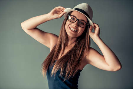 Beautiful woman in casual clothes, eyeglasses and stylish hat is posing at camera and smiling, on gray backgroundの写真素材