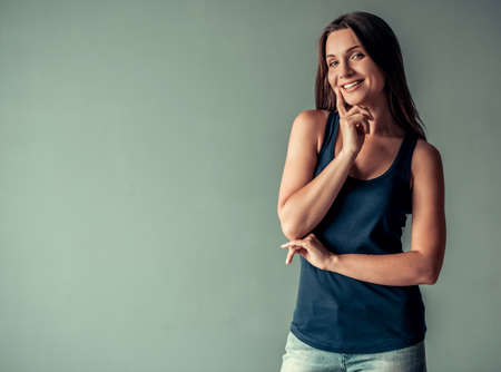 Beautiful woman in casual clothes is touching her lip, looking at camera and smiling, on gray backgroundの写真素材