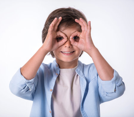Attractive little boy is making glasses, looking at camera and smiling, on light backgroundの写真素材