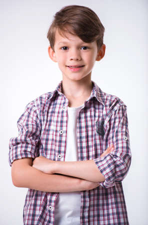 Handsome little boy is looking at camera and smiling while standing with crossed arms on light backgroundの写真素材