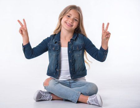 Beautiful little girl is showing peace signs, looking at camera and smiling while sitting on the floor on light backgroundの写真素材