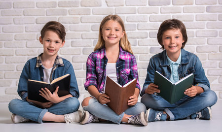 Cute kids are reading books, looking at camera and smiling while sitting on the floorの写真素材