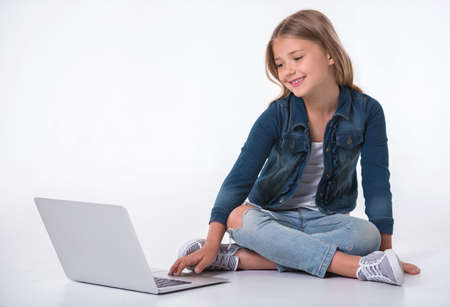 Beautiful little girl is using a laptop and smiling while sitting on the floor on light backgroundの写真素材