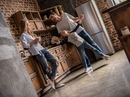Cute little girl and her handsome dad are dancing, mom is drinking tea, all are smiling while spending time together in kitchenの写真素材