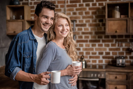 Beautiful young couple is holding cups, hugging and smiling while spending time together in kitchenの写真素材