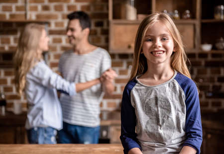 Cute little girl is looking at camera and smiling while her beautiful parents are dancing in the background in kitchenの写真素材