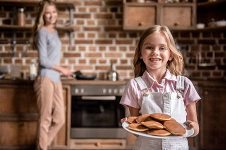 Cute little girl is holding pancakes, looking at camera and smiling while cooking together her beautiful mom in kitchenの写真素材