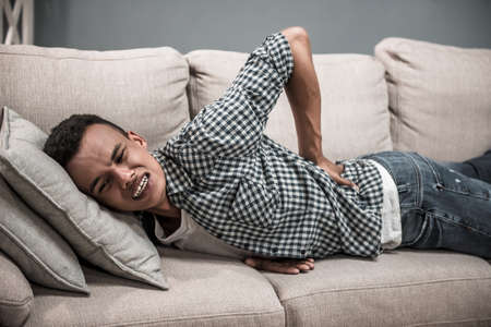 Attractive Afro American guy in casual clothes is touching his back feeling disastrous pain while lying on couch at homeの写真素材