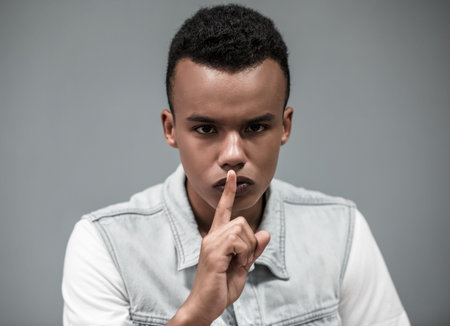 Attractive Afro American guy is showing silence sign and looking at camera, on gray background