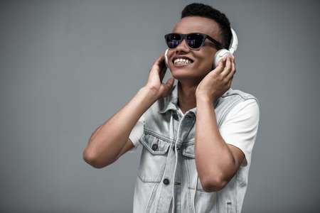 Stylish Afro American guy in headphones and sun glasses is listening to music and smiling, on gray backgroundの写真素材