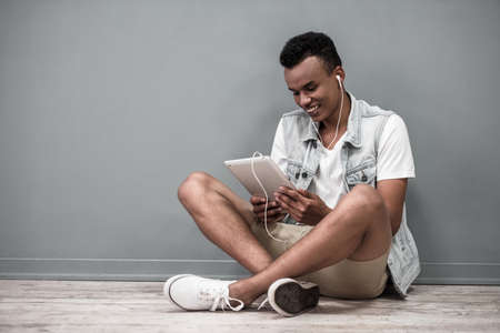 Stylish Afro American guy in earphones is using a digital tablet and smiling while sitting on the floorの写真素材