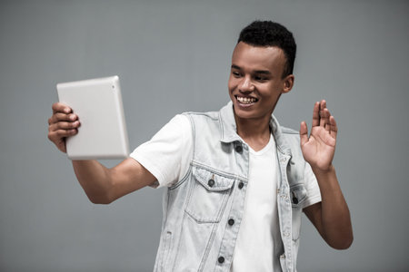 Attractive Afro American guy in stylish jean sleeveless jacket is using a digital tablet, waving and...