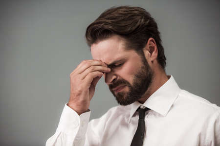 Handsome tired bearded businessman in formal suit is massaging his nose bridge, on gray backgroundの写真素材