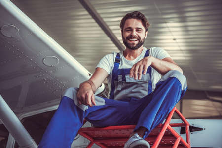 Handsome bearded mechanic in uniform is looking at camera and smiling while sitting near the aircraft in hangarの写真素材
