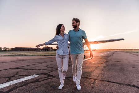 Beautiful romantic couple is hugging, looking at each other and smiling while walking on take-off ground near the aircraftの写真素材