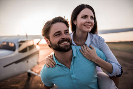 Beautiful romantic couple is looking away and smiling while standing on take-off ground near the aircraft, girl is piggybackの写真素材