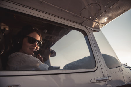 Beautiful woman in aviation headset is looking at camera and smiling while sitting in aircraft ready...