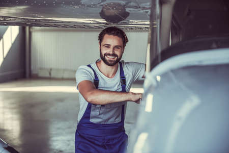Handsome bearded mechanic in uniform is looking at camera and smiling while opening an aircraft door in hangarの写真素材