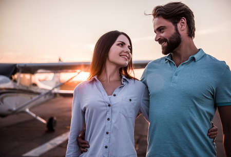 Beautiful romantic couple is hugging, looking at each other and smiling while walking on take-off ground near the aircraftの写真素材
