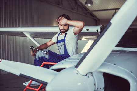 Handsome bearded mechanic in uniform is wiping his forehead while repairing an aircraft in hangarの写真素材