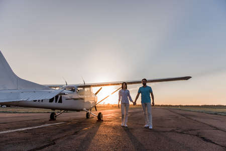 Beautiful romantic couple is holding hands, looking at camera and smiling while standing on take-off ground near the aircraftの写真素材
