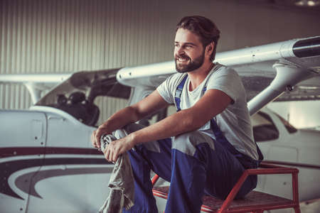 Handsome bearded mechanic in uniform is looking away and smiling while sitting near the aircraft in hangarの写真素材