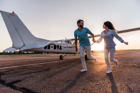 Beautiful romantic couple is holding hands, looking at each other and smiling while running on take-off ground near the aircraftの写真素材