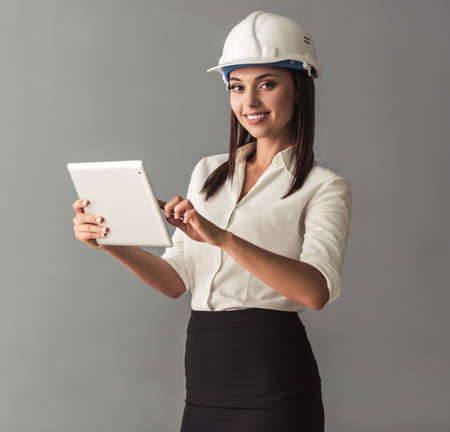Beautiful young business woman in suit and protective helmet is using a digital tablet, looking at camera and smiling, on gray backgroundの写真素材