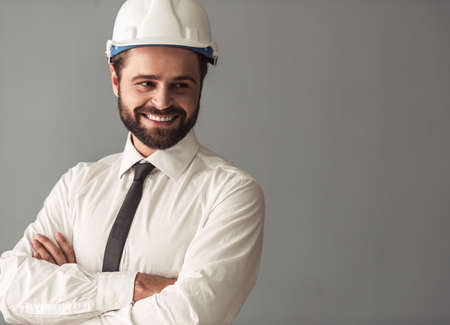Handsome bearded businessman in suit and protective helmet is looking away and smiling while standing with crossed arms on gray backgroundの写真素材