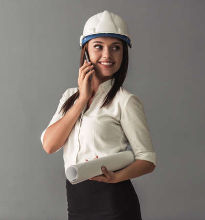 Beautiful young business woman in suit and protective helmet is talking on the mobile phone, holding rolled paper and smiling, on gray backgroundの写真素材