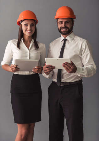 Handsome businessman and beautiful business woman in suits and protective helmets are using digital tablets, looking at camera and smiling, on gray backgroundの写真素材