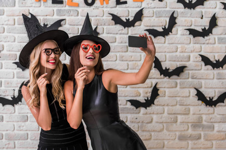 Beautiful girls in black dresses and witch hats are holding party props and doing selfie using a...