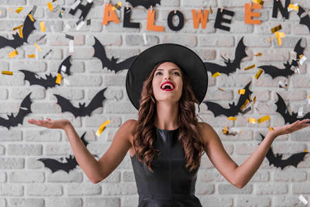 Beautiful girl in black dress and witch hat is looking upward being happy about confetti and smiling, on background decorated for Halloweenの写真素材