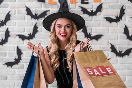 Beautiful blonde girl in black dress and witch hat is holding shopping bags, looking at camera and smiling, on background decorated for Halloweenの写真素材