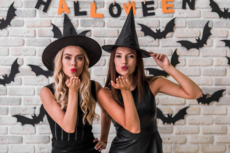 Beautiful girls in black dresses and witch hats are looking at camera and sending air kisses, on...