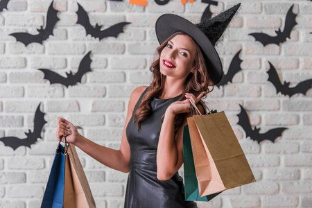 Beautiful girl in black dress and witch hat is holding shopping bags, looking at camera and smiling,...